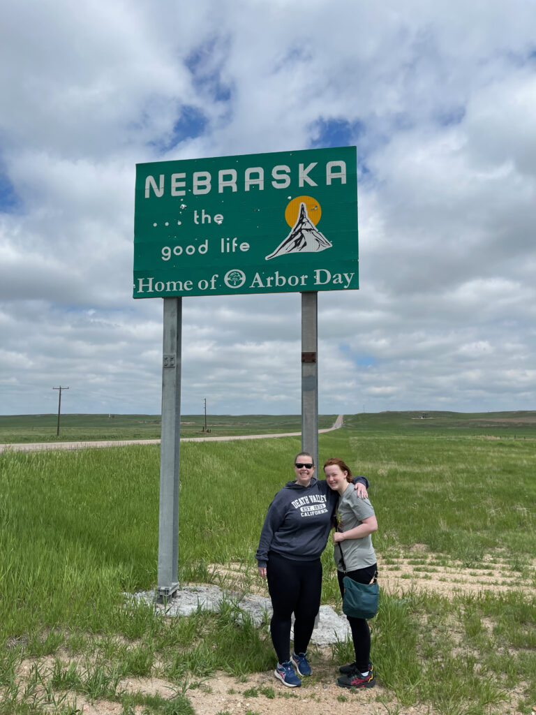 Cat with her arm around Cameron standing in front of the Nebraska the good life sign. Endless fields and a long road into the distance are in the background.