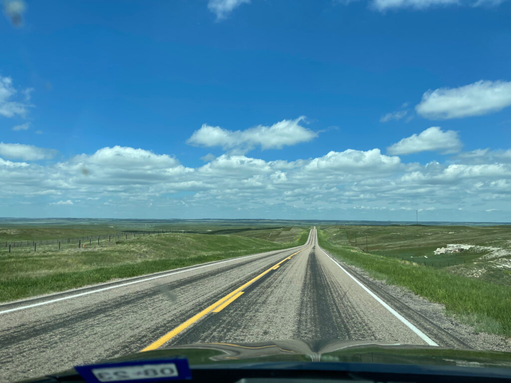 A view out the front of my car, as we drive down what looks like an endless two lane road through Nebraska. With a blue sky and a few clouds.