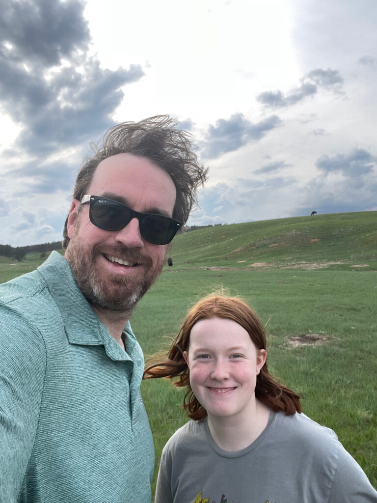 Cameron and myself wearing sunglasses stand new a Prairie Dog field with a lone Bison behind us on top of the hill in the distance. 