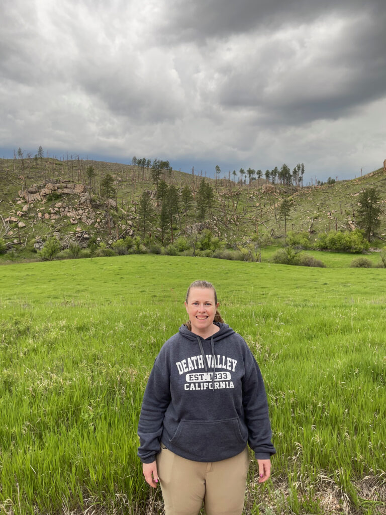 Cat wearing hiking pants and a Death Valley Sweatshirt standing in the middle of a field near Wind Cave National Park. Storm clouds are overhead. 