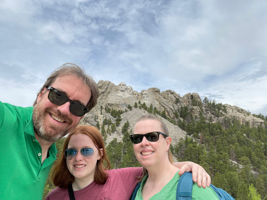 Cat, Cameron and myself standing in front of Mount Rushmore on a partly cloudy afternoon. All wearing t-shirts and sunglasses. 