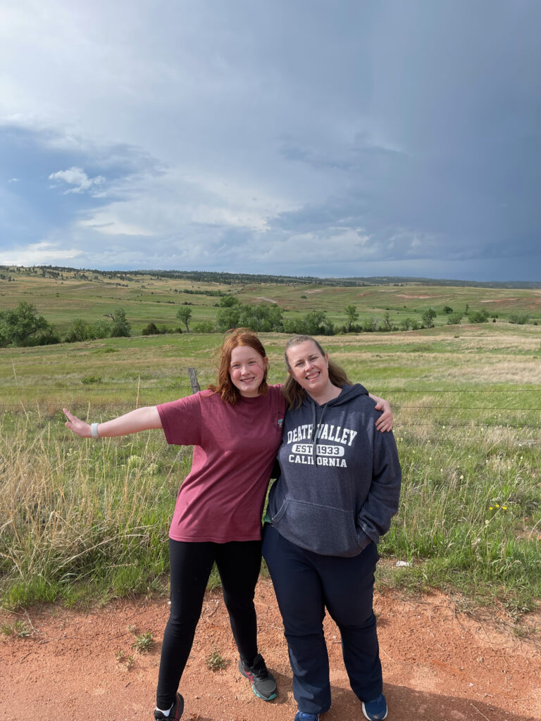 Cat and Cameron pose for a photo near a barbed wire fence, with storm clouds gathering over sprawling fields behind them.