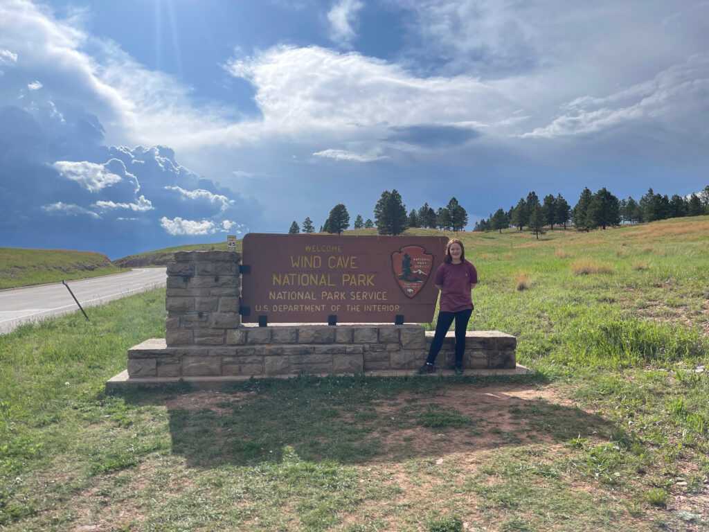 Cameron wearing a t-shirt standing next to the Wind Cave National Park sign. Storm clouds are overhead and the road into the the park is on the left. 