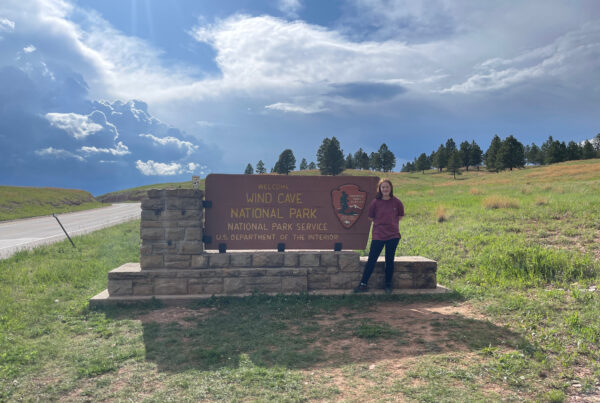 Cameron wearing a t-shirt standing next to the Wind Cave National Park sign. Storm clouds are overhead and the road into the the park is on the left.