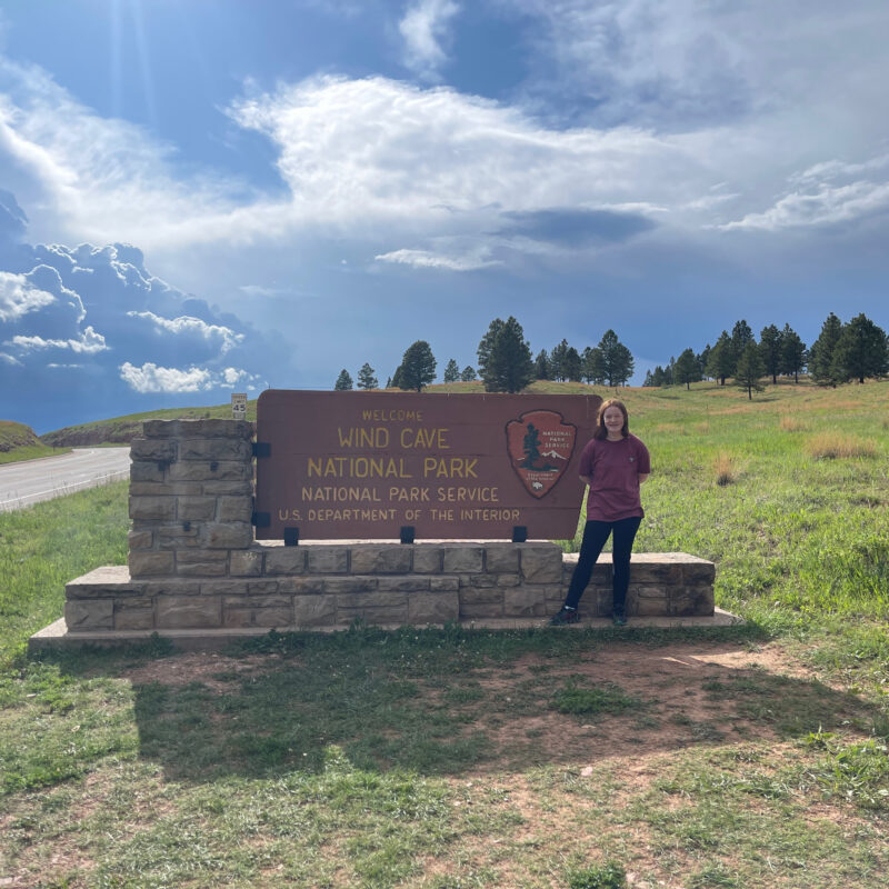 Cameron wearing a t-shirt standing next to the Wind Cave National Park sign. Storm clouds are overhead and the road into the the park is on the left.