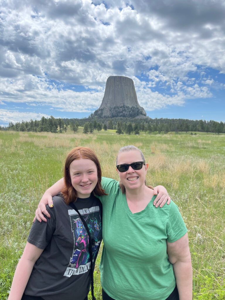 Cat and Cameron stand arm in arm in front of field and the Devils Tower behind them. Bright clouds are overhead. 