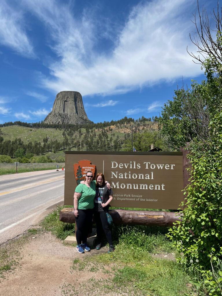 Cat and Cameron standing arm in arm, in front of the sign to the Devils Tower National Monument, with the road next to them and the tower in the background.