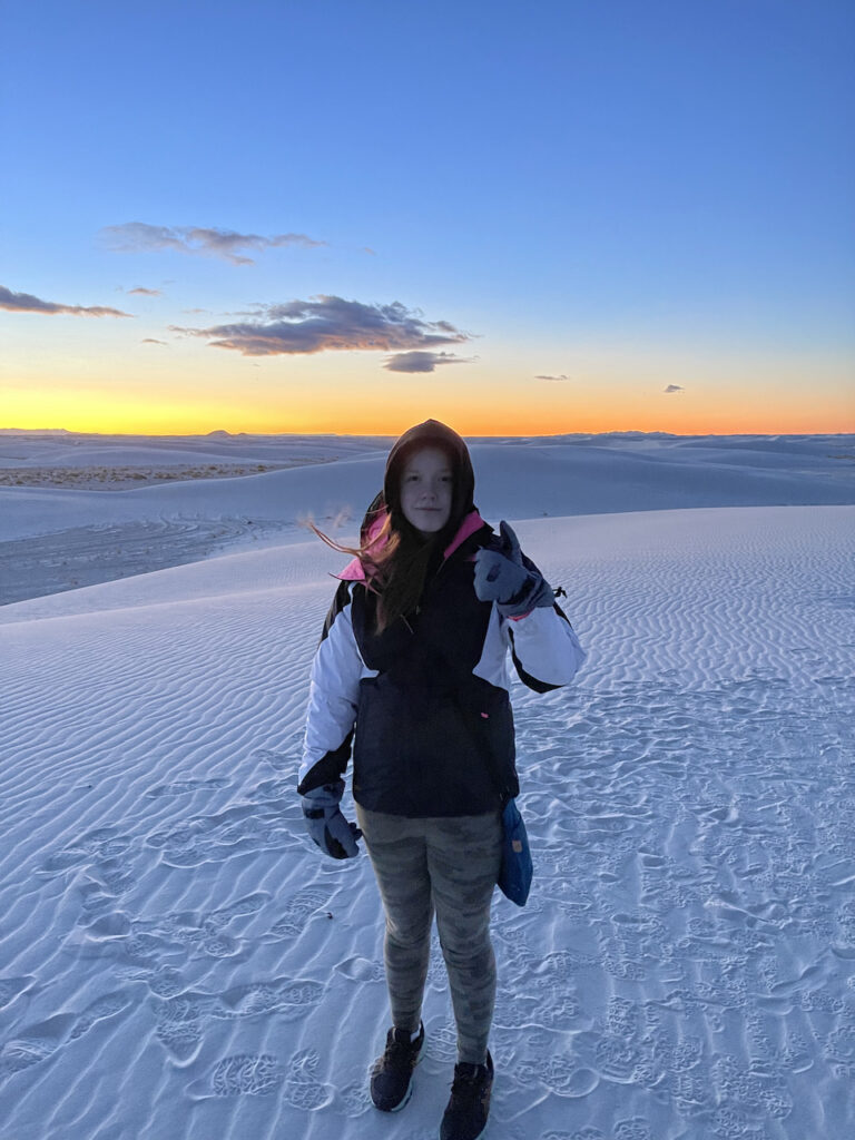 Cameron in a jacket, hat and gloves standing on the white sand in the pre-dawn light of White Sands National park. The sky is starting to turn yellow and orange and reflect on the sand.