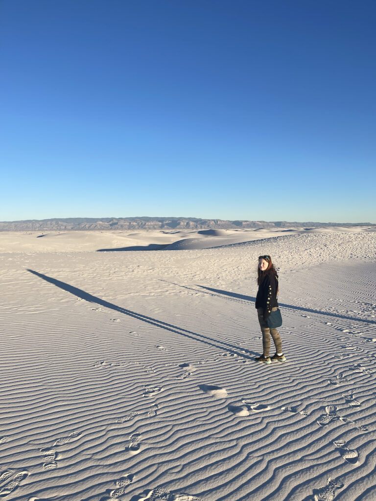 Cami standing alone on the White Sand dunes with her long shadow that reaches out in front of her. The sky is perfectly clear and deep blue.