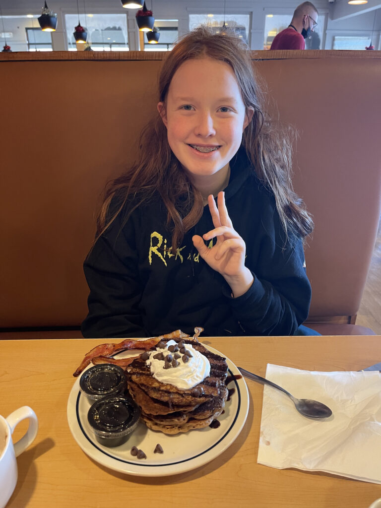 Cameron smiling and sitting in a booth at breakfast. With a massive plate of pancakes in front of her. 