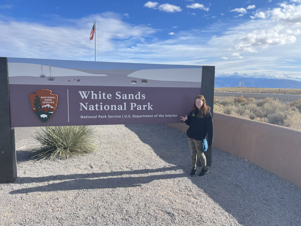 Cameron standing in front of the White Sands National Park sign, making a peace sign, with blue sky and clouds over the desert in the background.