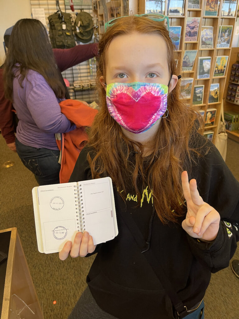 Cameron wearing a facemask, in the visitors center making a peace sign and showing her brand new passport stamp.