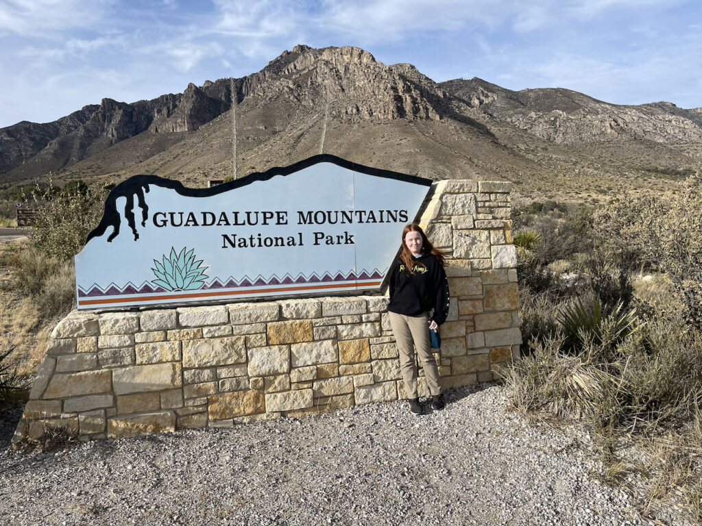 Cameron in front of the sign for the Guadalupe Mountains National Park with the massive parks mountains in the background.