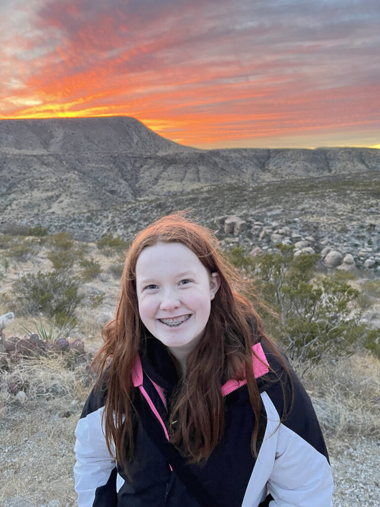 Cameron smiling on a trail in the Guadalupe Mountains. Her red hair is glowing from the sunset light along with the clouds in the sky above us.