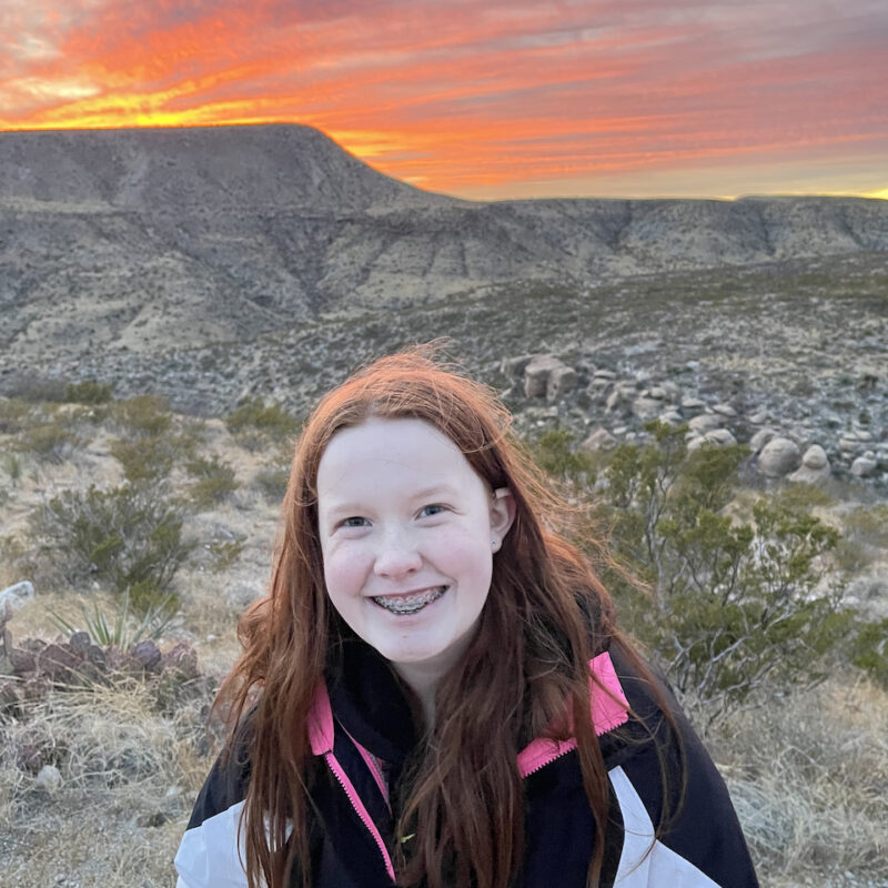 Cameron smiling on a trail in the Guadalupe Mountains. Her red hair is glowing from the sunset light along with the clouds in the sky above us.