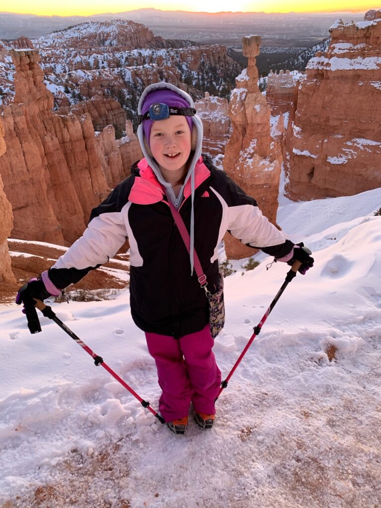 Cameron standing in the early morning on the Navajo Loop Trial, it is cover with thick ice and Cami has her hand lamp, winter coat, trekking polls and crampons. 