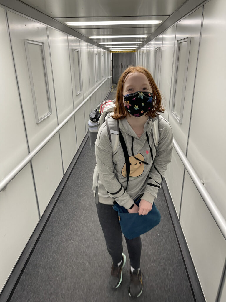 Cameron carrying her backpack and purse, wearing a face mask alone on the jet-bridge about to board a flight. 