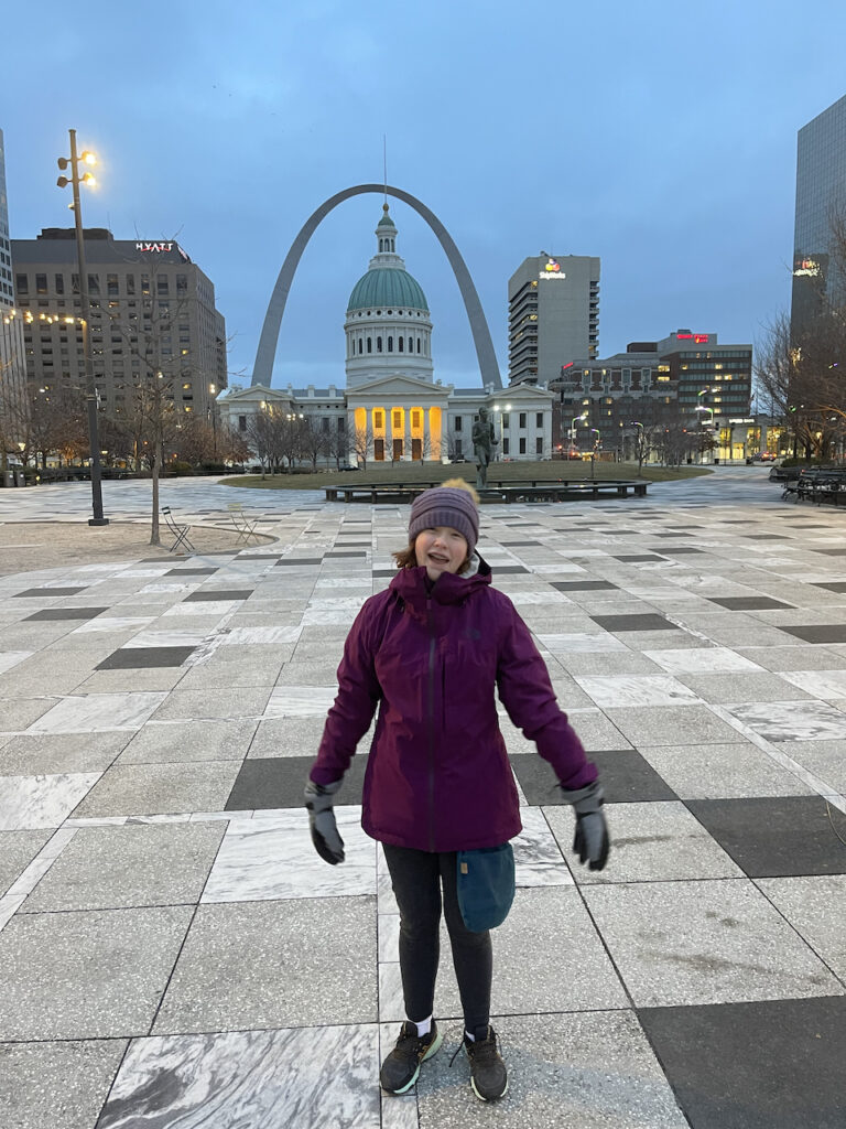 Cameron at sunrise in a hat, gloves and hat, in the pre-dawn hours in Kiener Plaza with the Gateway Arch and State house in the background.