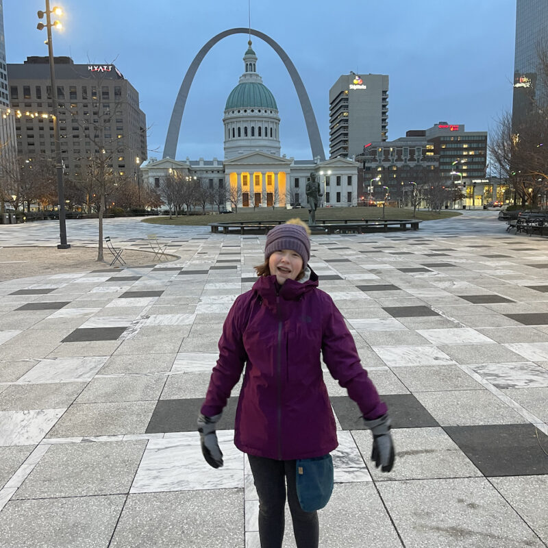 Cameron at sunrise in a hat, gloves and hat, in the pre-dawn hours in Kiener Plaza with the Gateway Arch and State house in the background.