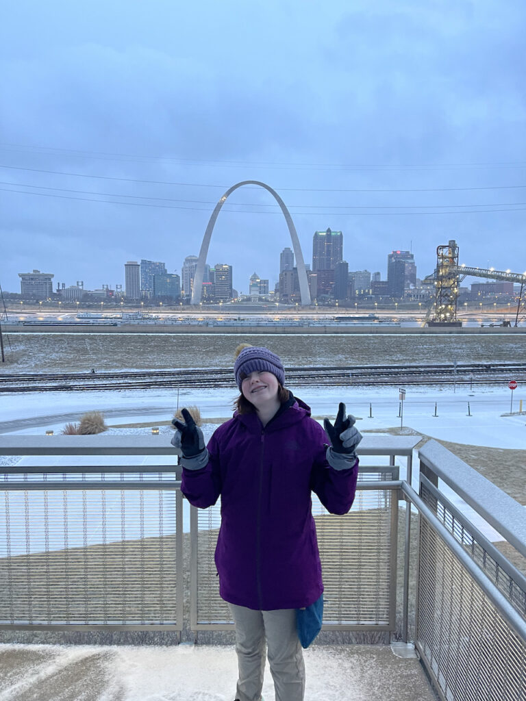 Cameron in East St Louis at sunset, in the winter with hat, gloves and a big winter jacket. Behind her are the train track, the Mississippi river and the Gateway Arch.