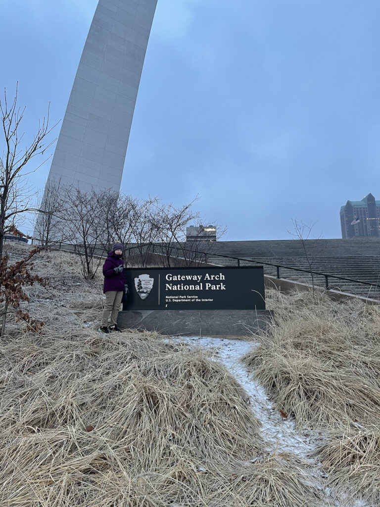 Cameron on a cold and early winters morning, wearing a jacket, hat and gloves, standing by the snow by the Gateway Arch National Park sign.