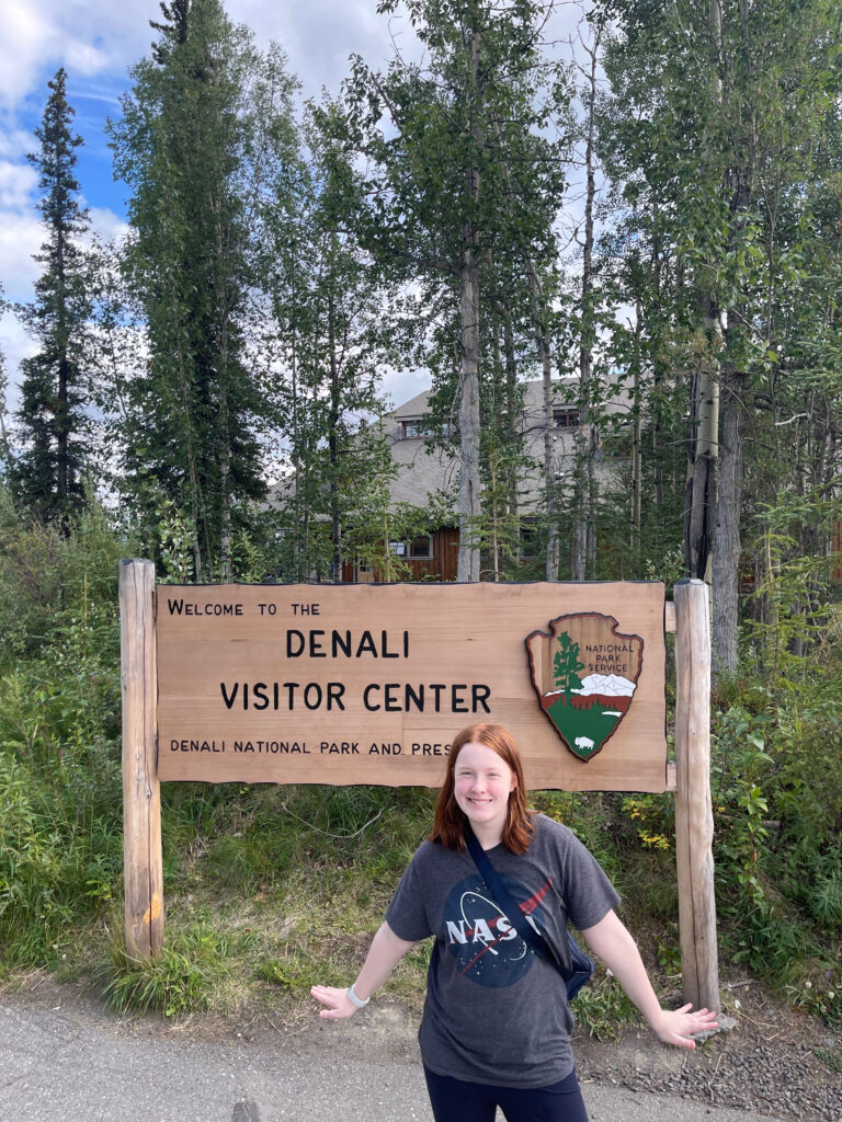 Cameron posing next to the Denali Visitors Center wooden sign, in the middle of a bright day.