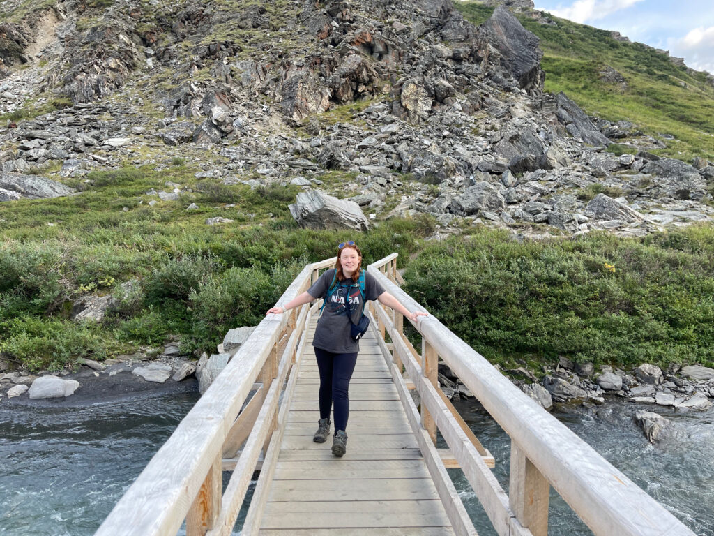 Cameron standing on the middle of a wooden bridge, both hands on the railing, wearing hiking gear and a backpack. Taken on the Savage Alpine trail in Denali.