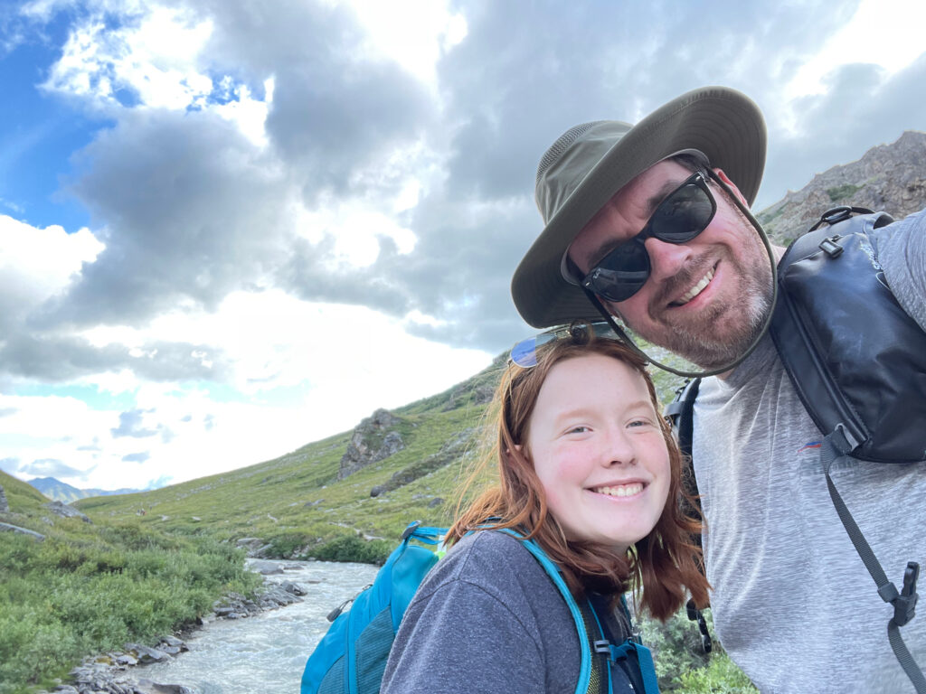 A photo of Cameron and myself - taken on the Savage Alpine Trail inside Denali. Both wearing hiking clothing and me with a hate and sunglasses on. The Savage river is next to us and the sky is full of some white clouds and blue sky.