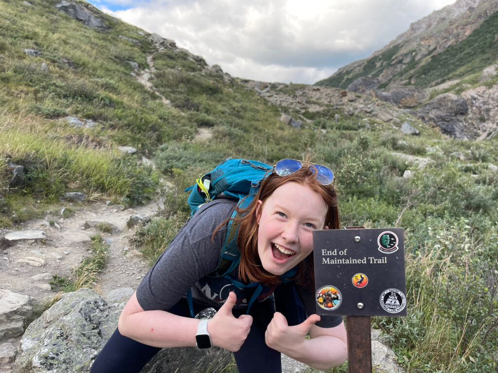 Cameron squatting down with a huge smile and two thumbs on, on  the trail in Denali next to a sign that says "End of Maintained Trail"