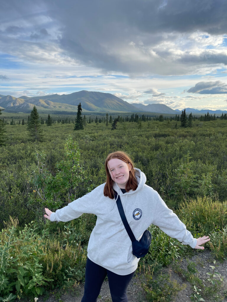 Cameron posing in a massive open grass land in Denali, mountains and clouds can be seen in the distance. 