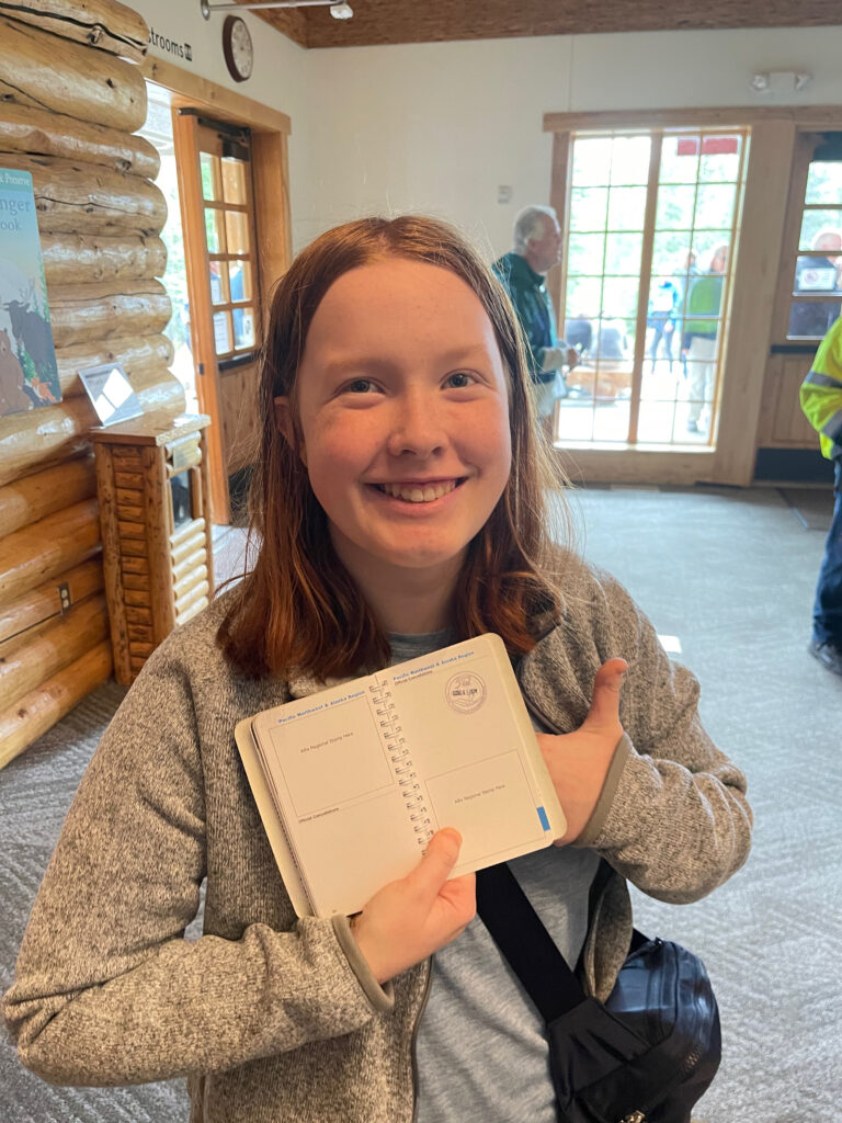 Cameron standing inside the visitors center at Denali National Park. Holding open her National Park Passport book to show the stamp and giving a thumbs up she is all smiles. 