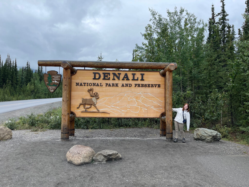 Cameron standing next to and holding onto the massive Denali National Park and Preserve sign by the side of the road on a cloudy day.