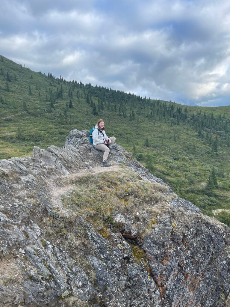 Cameron in hiking cloths, a backpack and camera bag, sitting on rocks high up on the Savage Alpine Trail.