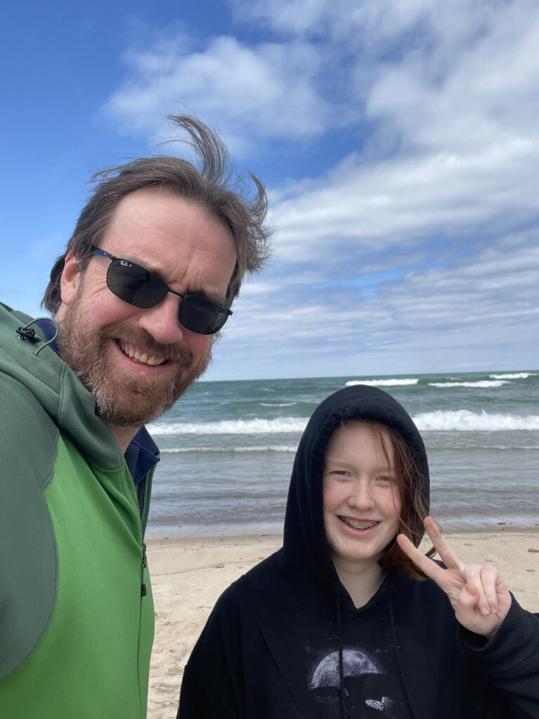 Cameron and myself on the beach, smiling and cami making a peace sign. The lake is behind us with white puffy clouds.