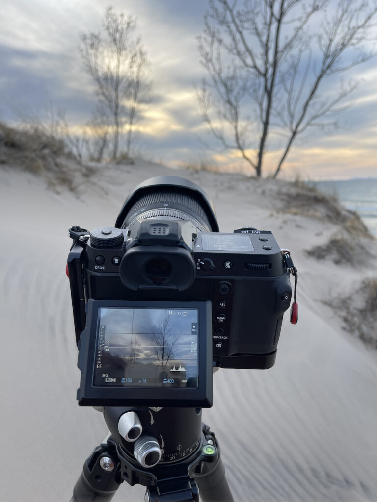 My camera on a tripod, halfway up a dune, pointed at the trees on the edge of the dune an the cloudy sky that is just starting to get some sunset color.