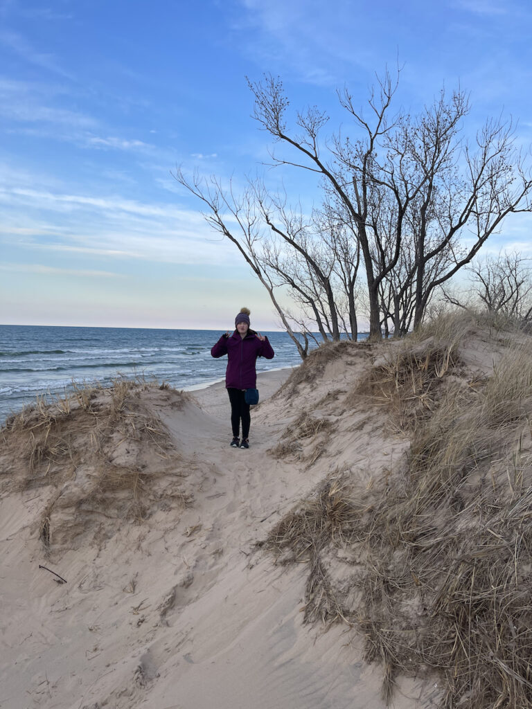 Cameron walking on a trial near the tops of the dunes, with some grass and trees around her and the lake and big almost clear sky behind her.