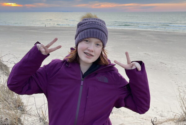 Cameron wearing a light jacket, hat, and making a double peace sign, on top of the Dunes at the National Park. The sky is full of clouds but has a hint of yellow and orange color above the calm lake.