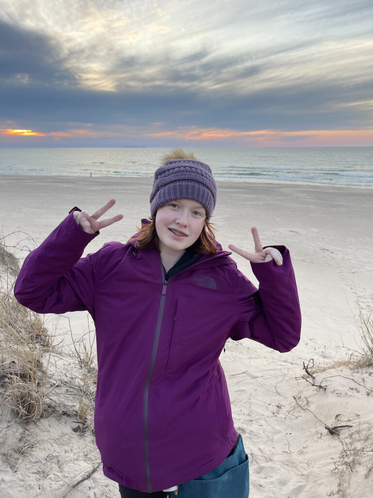 Cameron wearing a light jacket, hat, and making a double peace sign, on top of the Dunes at the National Park. The sky is full of clouds but has a hint of yellow and orange color above the calm lake.