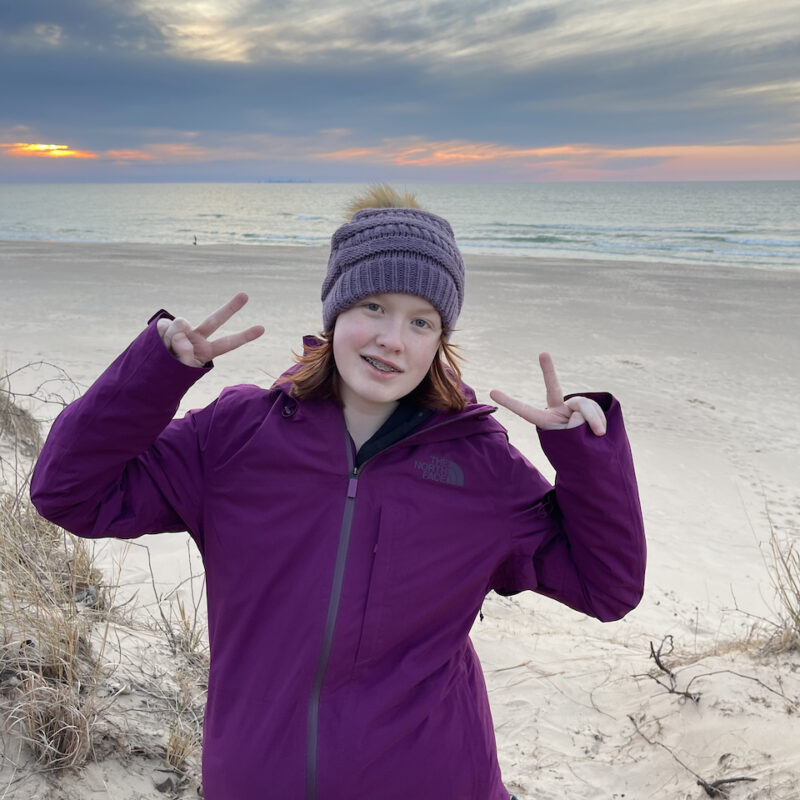 Cameron wearing a light jacket, hat, and making a double peace sign, on top of the Dunes at the National Park. The sky is full of clouds but has a hint of yellow and orange color above the calm lake.