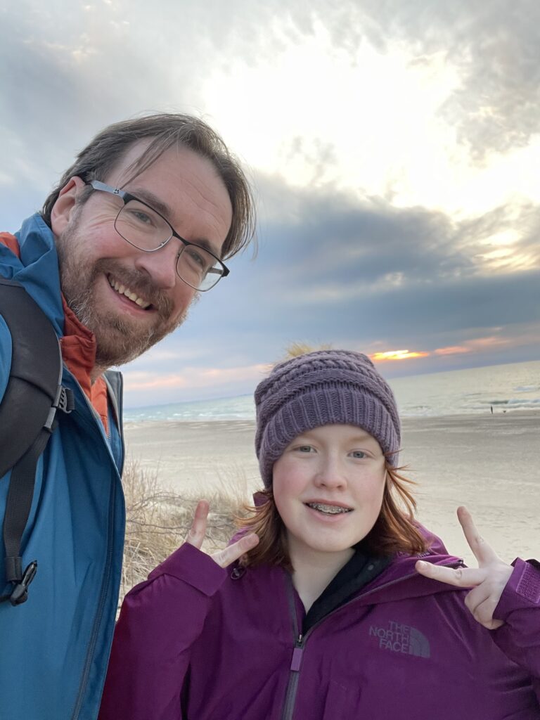 Cameron and myself at sunset, both wearing jackets and smiling, Cami is making a peace sign. A sky full of colorful clouds and the lake are behind us.