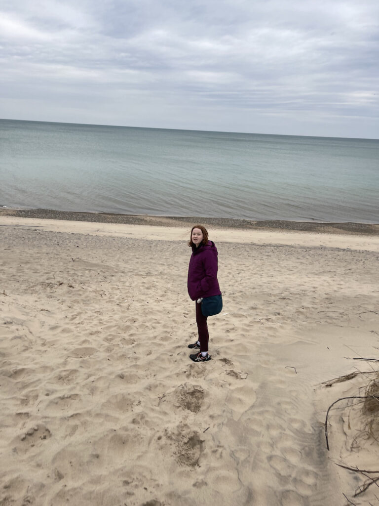 Cameron wearing a purple jacket, walking down the beach, leaving footprints behind her. The very calm lake is in front of her.