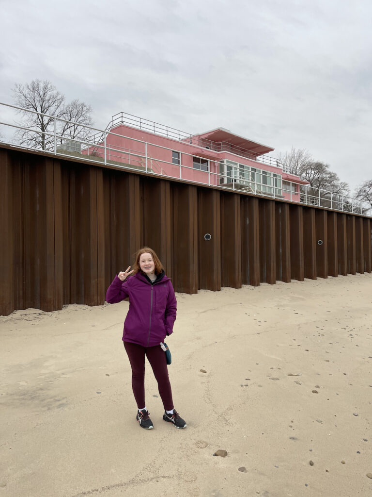 Cameron smiling on the beach with a massive sea wall behind her. On top is the pink Florida Tropical House built in 1933.