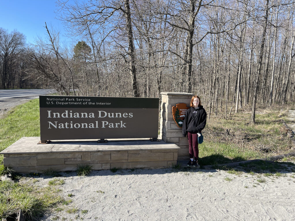 Cameron in front of the park sign for Indiana Dunes National Park. In winter with bare trees and the road in the background.