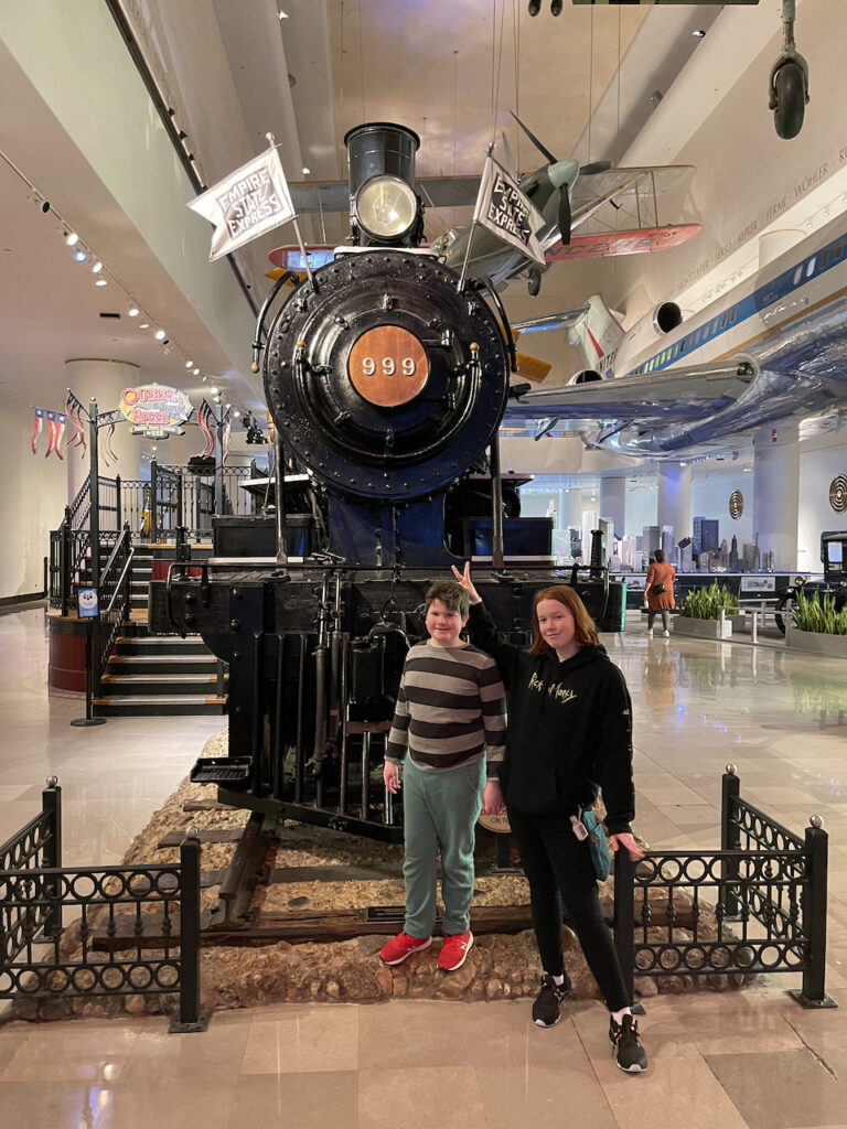 Cameron and Collin in front of a massive old steam train, inside the Museum of Science and Industry. Cami is making buddy ears behind collins head and both kids are all smiles. 