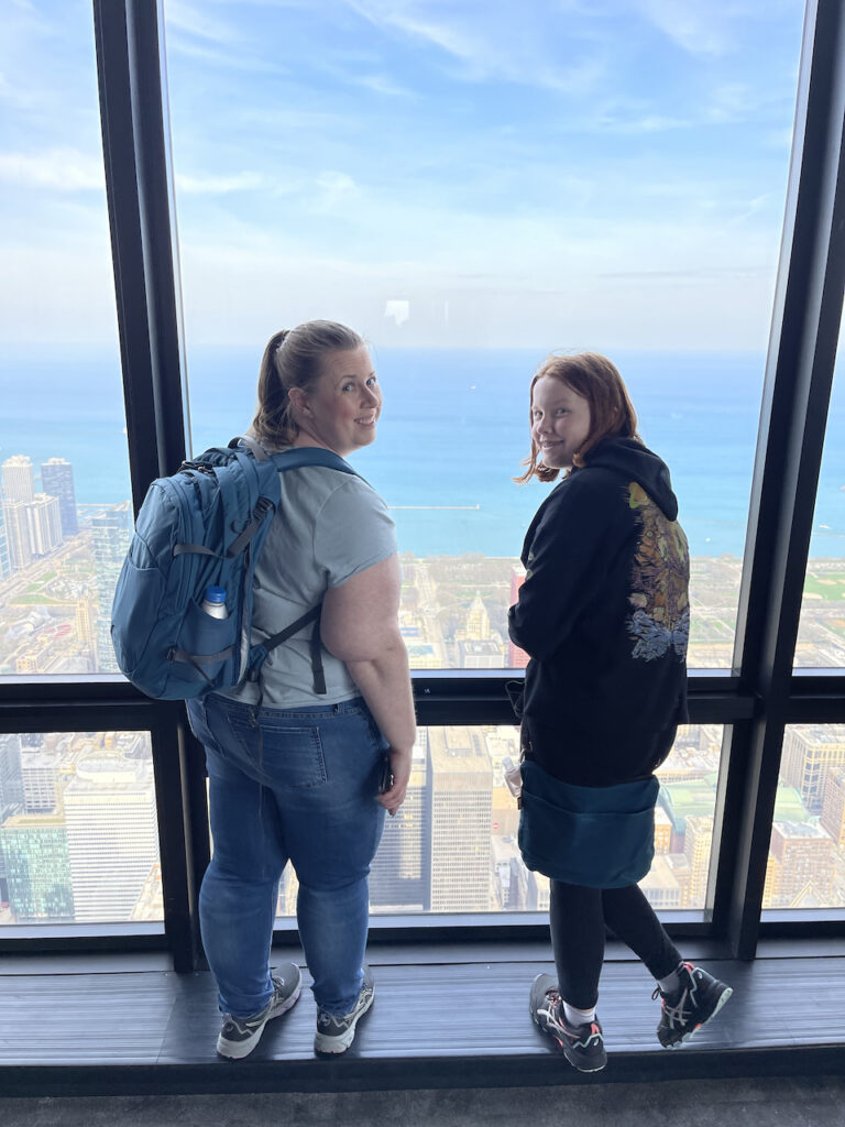 Cat wearing her backpack standing next to Cameron in front of the Windows looking down at the city of Chicago from the Skydeck of the Willis Tower.