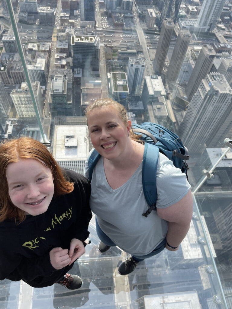 Cat and Cameron looking up the camera in one of the all glasses boxes that hangs over the edge of the Willis Tower, on the Skydeck looking down at the city of Chicago.