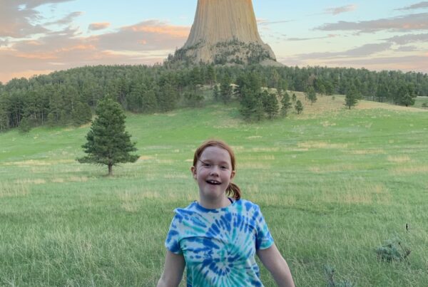 Cameron posing at sunset on the Joyner Ridge Trail, in a grassy filed. The last bit of sunlight is hitting the top of the Devils Tower and the sky is full of colorful clouds.