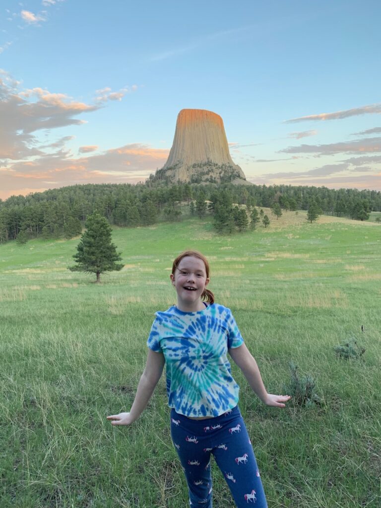 Cameron posing at sunset on the Joyner Ridge Trail, in a grassy filed. The last bit of sunlight is hitting the top of the Devils Tower and the sky is full of colorful clouds. 