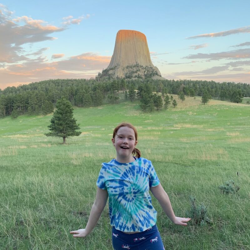 Cameron posing at sunset on the Joyner Ridge Trail, in a grassy filed. The last bit of sunlight is hitting the top of the Devils Tower and the sky is full of colorful clouds.