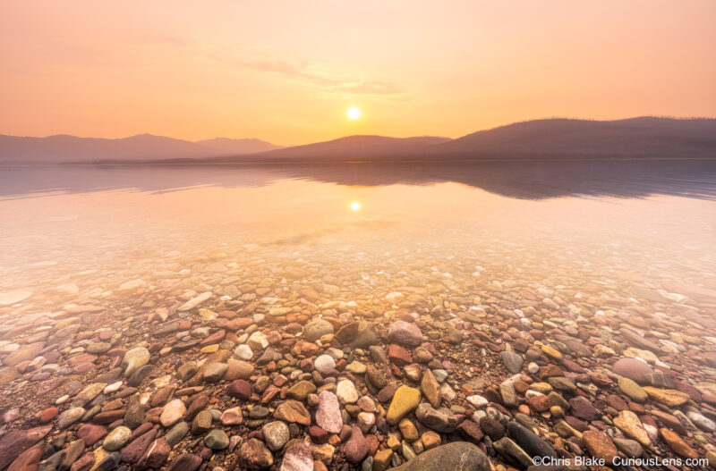 Lake McDonald in Glacier National Park with smoky conditions, soft yellow filter effect, sunset reflection, and rays of light on the lake.